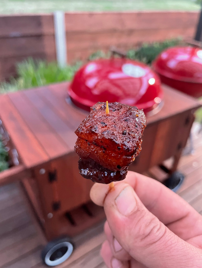 Glazed pork belly skewer held up in front of red Weber kettles