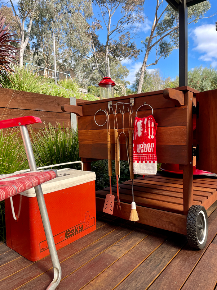 Weber tool cart with utensils and a red Esky cooler on a timber deck