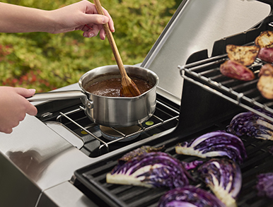Sauce being stirred on the side burner next to grilled potatoes and cabbage