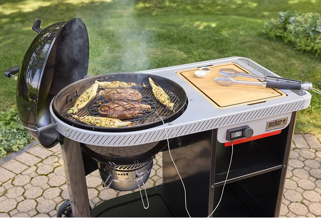 Weber Performer charcoal barbecue with meat and vegetables grilling on the cooking grate and tongs resting on the side table.